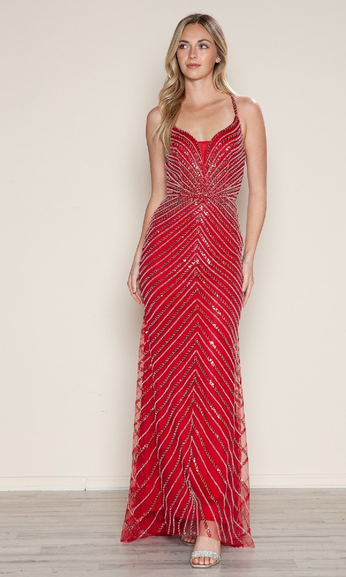 A woman poses by a plain wall wearing the Cut-Out Long Beaded Prom Dress: Poly USA 9530, a floor-length fitted red gown with silver sequin designs and spaghetti straps. She pairs it with open-toe heels, ideal for formal events.